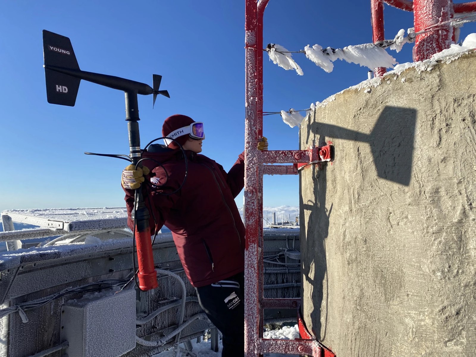 A Mount Washington Observatory staffer on the icy summit tower holding a wind vane
